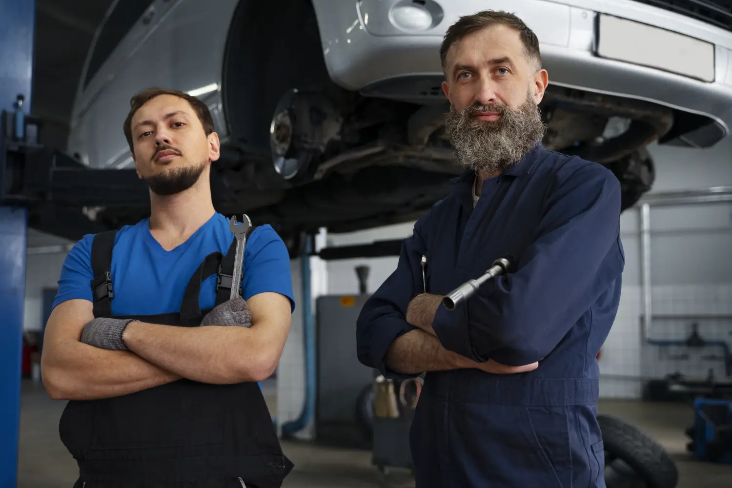 Vr-Transport - Transport · Reifen & KFZ-Ersatzteile - Zwei männliche Automechaniker, einer davon mit Bart, stehen selbstsicher mit verschränkten Armen in einer Autowerkstatt. Hinter ihnen wird ein silberner Wagen auf einer Hebebühne angehoben.